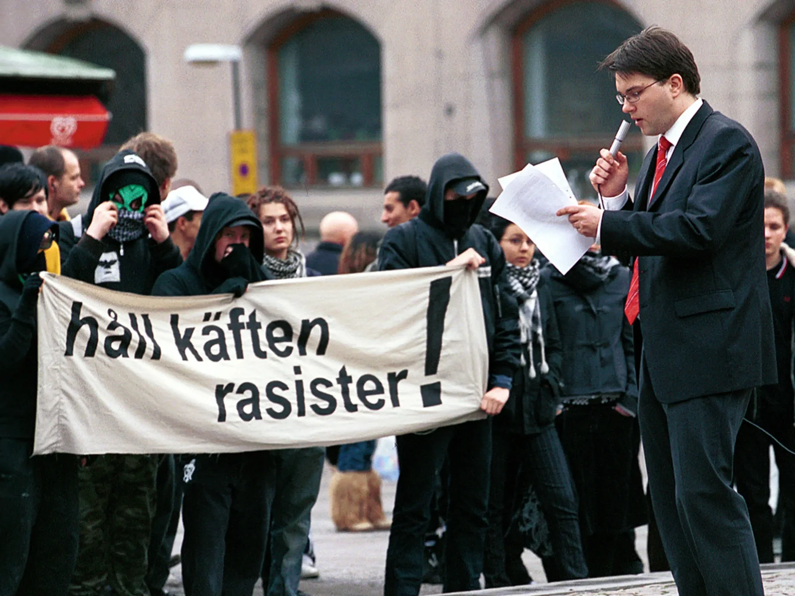 Jimmie Åkesson står och håller tal ur några papper, framför honom motdemonstranter med en banderoll: "Håll käften rasister!"